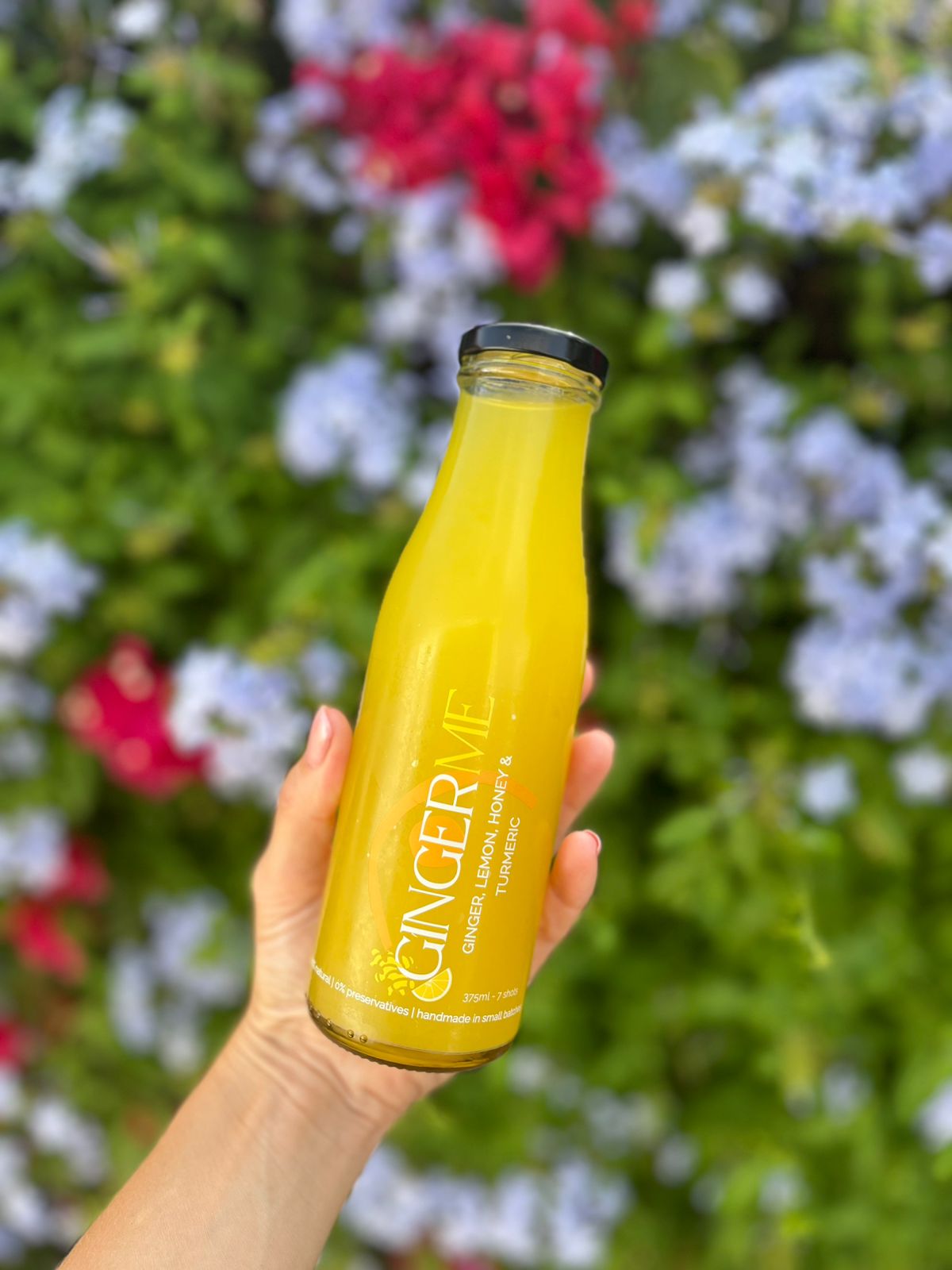 Hand holding a bottle of 'Ginger Me' juice with a blurred floral background