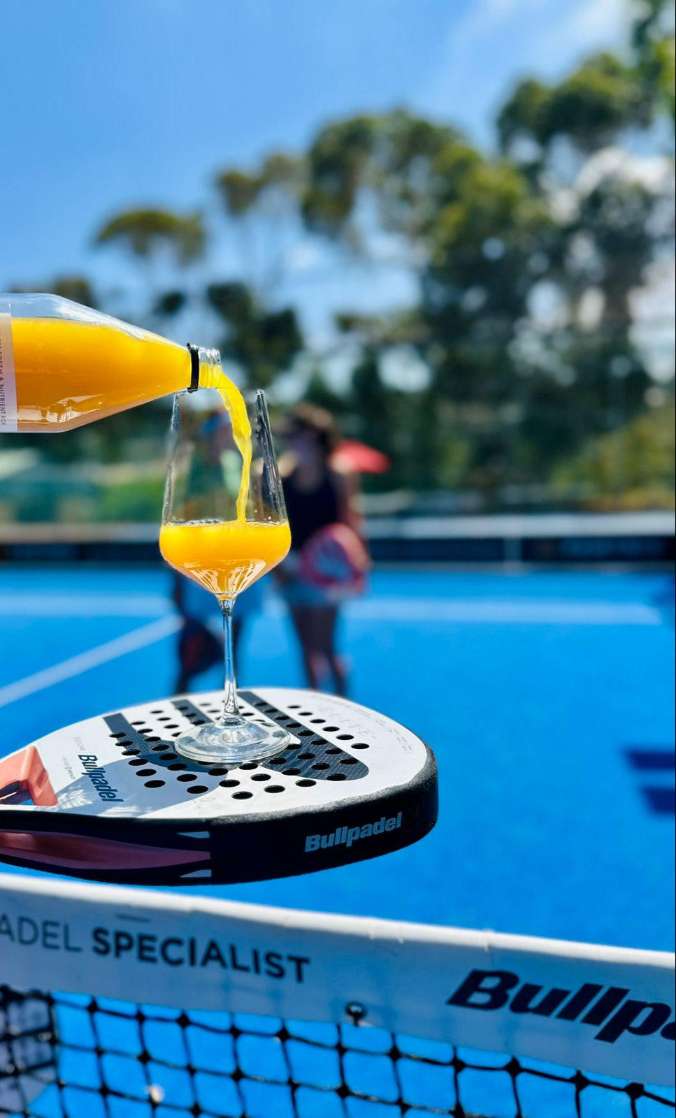 Orange ginger shots being poured into a glass on a paddle racket with a court and trees in the background