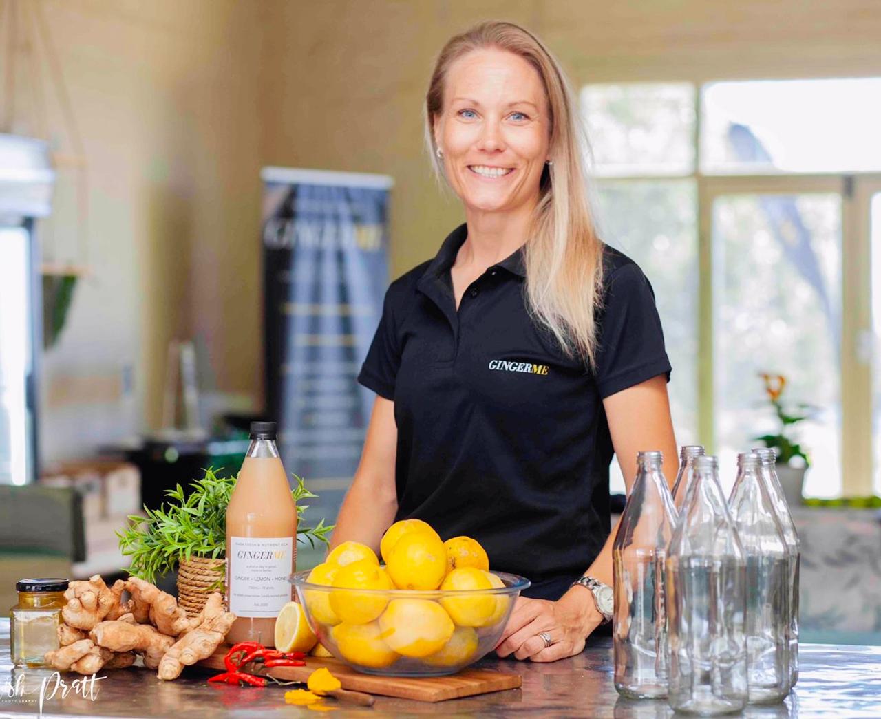 Woman in a black shirt with 'GingerMe' logo standing behind a table with lemons, ginger, and bottles.