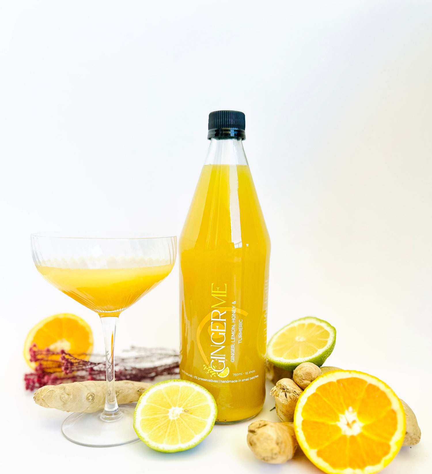 Bottle of 'GINGER ME' ginger juice with a glass of juice, lemons, and ginger roots on a white background