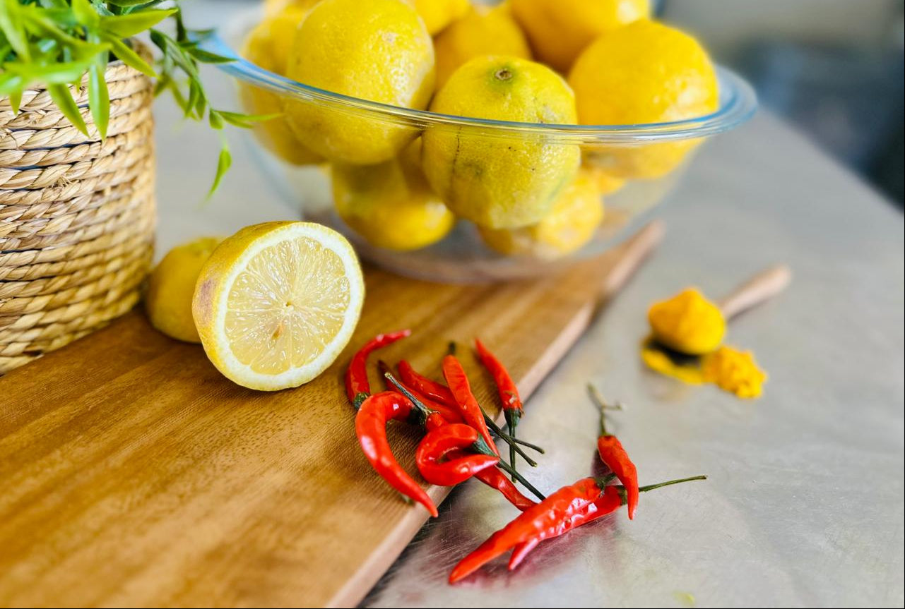 Glass bowl of lemons on a wooden surface with red chilli's and a plant.