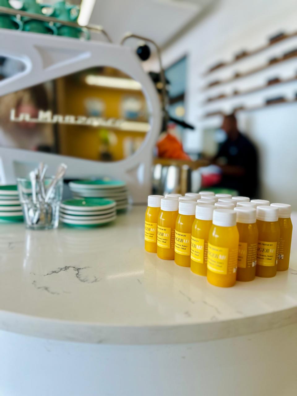 Row of 50ml 'GingerMe' shot bottles with white caps on a marble surface, with a blurred background.